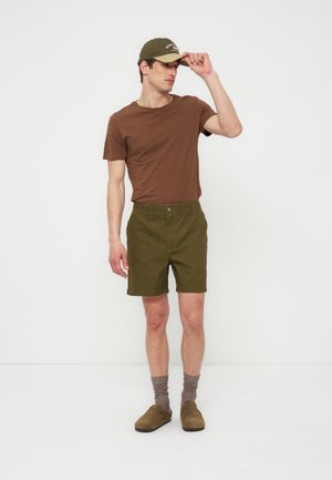 Muškarac u smeđoj majici, maslinasto zelenim kratkim hlačama, usklađenom kapom, sivim čarapama i smeđim kliznim cipelama, stoji nasuprot jednobojne bijele pozadine.