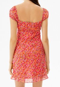 Vaaleanpunainen kukkamekko, jossa on lyhyet puffihihat, neliön muotoinen selkä ja röyhelöhelma. Kangas esittelee värikkään kukkakuosin ja kevyen rakenteen.