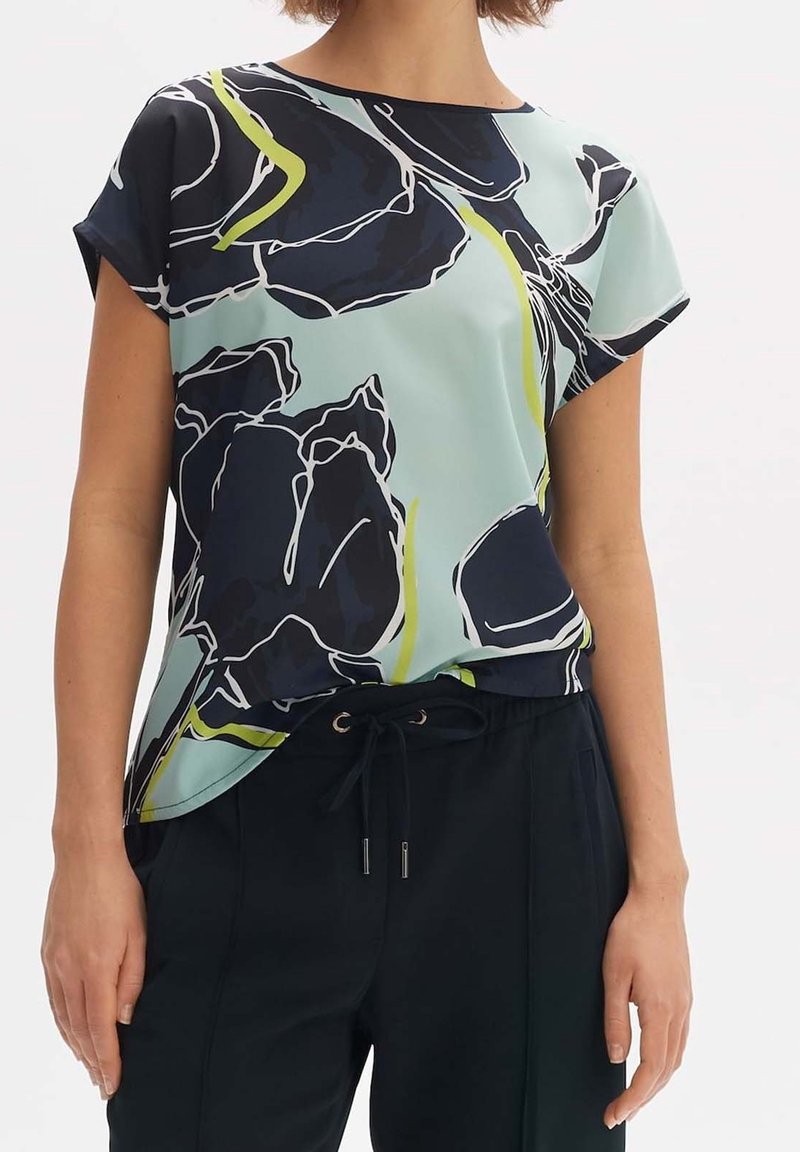 Krátký rukáv, světle modrý top s černým květinovým vzorem a zelenými akcenty. Vyrobený z hladké látky s volným střihem a zaobleným výstřihem.