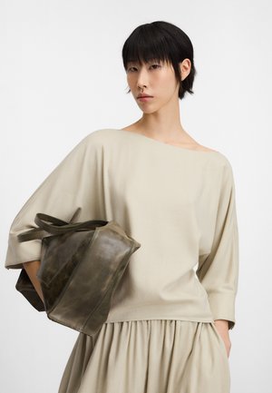 Kobieta o krótkich czarnych włosach nosi luźną beżową bluzkę i spódnicę, trzymając dużą ciemnooliwkową skórzaną torbę pod pachą na jednolitym tle.