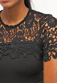 Svart klänning med intrikata svart spetsdetaljer vid halsen och ärmarna. Slät tyg med blommotiv och en figurnära silhuett.