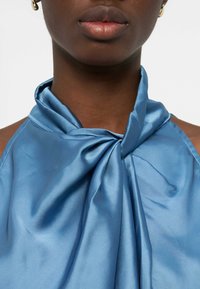 Gros plan sur une personne portant un haut dos nu en satin bleu brillant noué de manière lâche au niveau du col, avec de petites boucles d'oreilles en or.