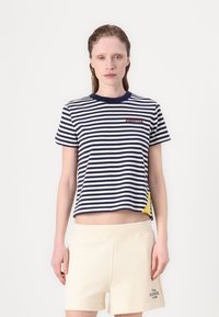 Jūrų ir baltos juostelės trumpa marškinėliai su(apvaliu) apykakle ir raudonu "HILFIGER" logotipu, derinami su šviesiais kreminiais šortais.