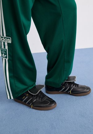 Persoon draagt groene Adidas trainingsbroek en zwarte Adidas Samba sneakers met bruine zolen op een lichtblauwe vloer.