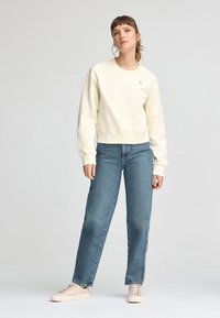Sudadera crema con cuello redondo y puños de canalé, combinada con jeans de mezclilla azul claro de pierna recta y zapatillas beige. Diseño sencillo.