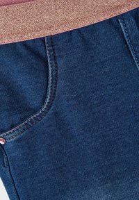 Denim-kangas tummansinisen sävyisenä, jossa on etutasku punaisella ompeleella ja hienovaraisella kiillolla, jonka päällä on tekstuurinen vaaleanpunainen vyötärönauha.