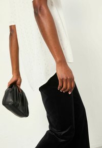 Weißes, strukturiertes Oberteil mit kurzen Ärmeln, kombiniert mit schwarzen, samtigen, weit geschnittenen Hosen. Die Hand hält eine zerknüllte schwarze Leder-Clutch.