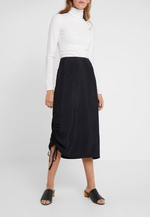 Falda midi de satén negra con fruncido lateral y detalle de lazo. Combinada con un suéter blanco de cuello alto y sandalias negras tipo slide con acentos amarillos.
