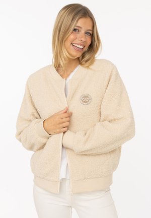 Lächelnde Frau trägt eine beige Sherpa-Jacke mit einem "Good Vibes Only" Herzlogo, halb geöffnet, steht vor einem weißen Hintergrund.