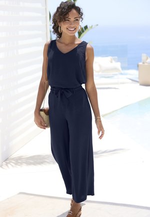 Navy blue jumpsuit with a V-neck, belted waist, and wide legs. Worn with brown sandals and a woven purse. Curly hair and hoop earrings.