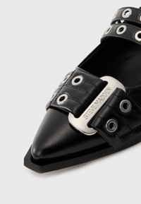 Black pointed-toe shoe with silver grommets and a metal buckle engraved "Steve Madden" on the strap across the front.