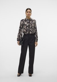 Zwarte blouse met lange mouwen en een wit bloemenpatroon, gecombineerd met zwarte op maat gemaakte broek. Accessoire: zwarte clutch tas. Schoeisel: zwarte ballerina's.