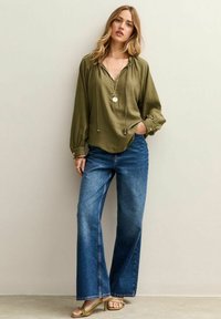 Olijfgroene blouse met lange mouwen, een V-hals en een trekkoord, gecombineerd met wijde blauwe spijkerbroeken voor een ontspannen pasvorm. Gouden sandalen.