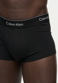 Κοντινό πλάνο ενός ατόμου που φοράει μαύρα μποξεράκια Calvin Klein με επώνυμη ζώνη μέσης σε απλό ανοιχτό φόντο.