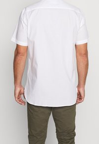 Uomo che indossa una camicia bianca a maniche corte e pantaloni verde oliva, visto da dietro su uno sfondo leggero e uniforme.