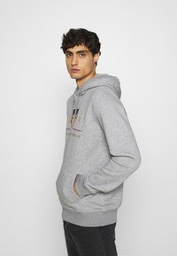 Grauer Kapuzenpullover mit einer vorderen Tasche und gesticktem Logo in Rot, Blau und Gold. Weicher Stoff mit gerippten Bündchen.