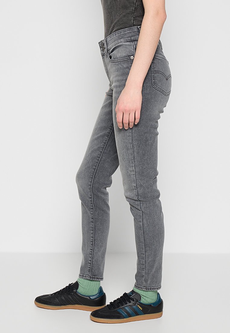 Persona vistiendo jeans ajustados de color gris desgastado, calcetines verdes y zapatillas negras con rayas azules, de pie contra una pared blanca lisa.