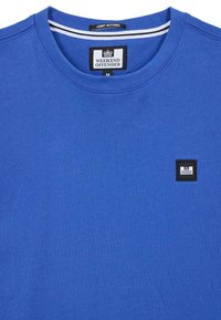 Blå bomull T-skjorte med rund hals og svart firkantet logo-patch på venstre bryst. Har horisontale stripe detaljer inni kragen.