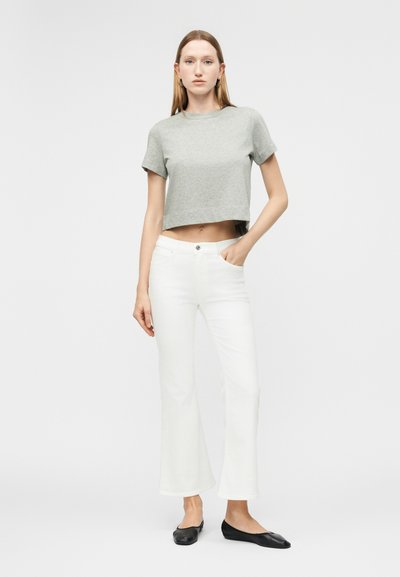 Γυναίκα όρθια που φοράει γκρι κοντό t-shirt, λευκά φαρδιά τζιν, μαύρες μπαλαρίνες, με το ένα χέρι στην τσέπη, μπροστά σε λευκό, μονόχρωμο φόντο.