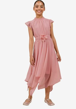Vestido midi de un suave color rosa pálido con un cuello alto, mangas cortas con volantes, cintura ceñida y un adorno de flor; dobladillo asimétrico que fluye. Zapatos planos marrones.