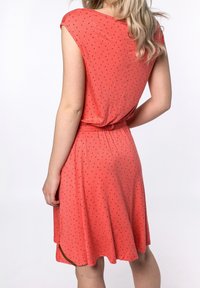 Korálové šaty s krátkým rukávem, elastickým pasem a černými srdcovými vzory. Měkká tkanina s rozšířenou sukní a bočními rozparky.
