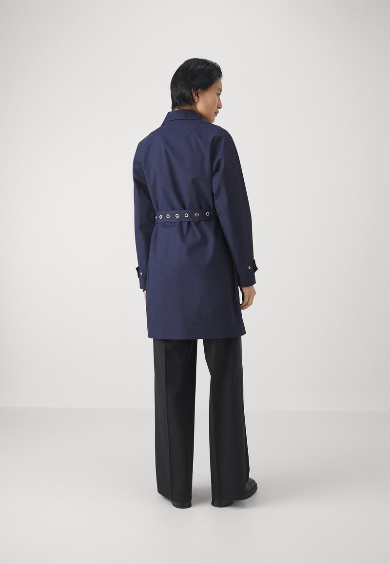 Abrigo azul marino con cintura ceñida por un cinturón, que presenta ojales dorados en el cinturón y mangas largas, combinado con pantalones de pierna ancha negros.