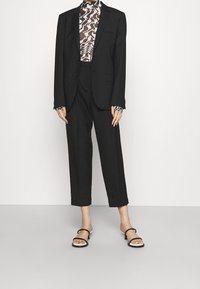 Persona che indossa un blazer nero su misura e pantaloni neri cropped a vita alta con una maglietta a maniche lunghe a fantasia astratta e tacchi neri con cinturini.