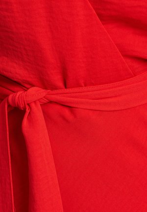 Crvena haljina s omotom i remenom, s glatkom teksturom i lepršavim materijalom. Dizajn uključuje duboki V-izrez i punu suknju.