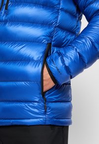 Veste matelassée bleue avec un design en coussins, poches zippées et un poignet élastique. Le matériau a une texture lisse et brillante. Marque "OMNI-HEAT" visible.