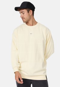 Sudadera de color crema con corte relajado, puños y dobladillo acanalados, y un pequeño logo de la marca en la parte delantera. Combinada con una gorra negra.