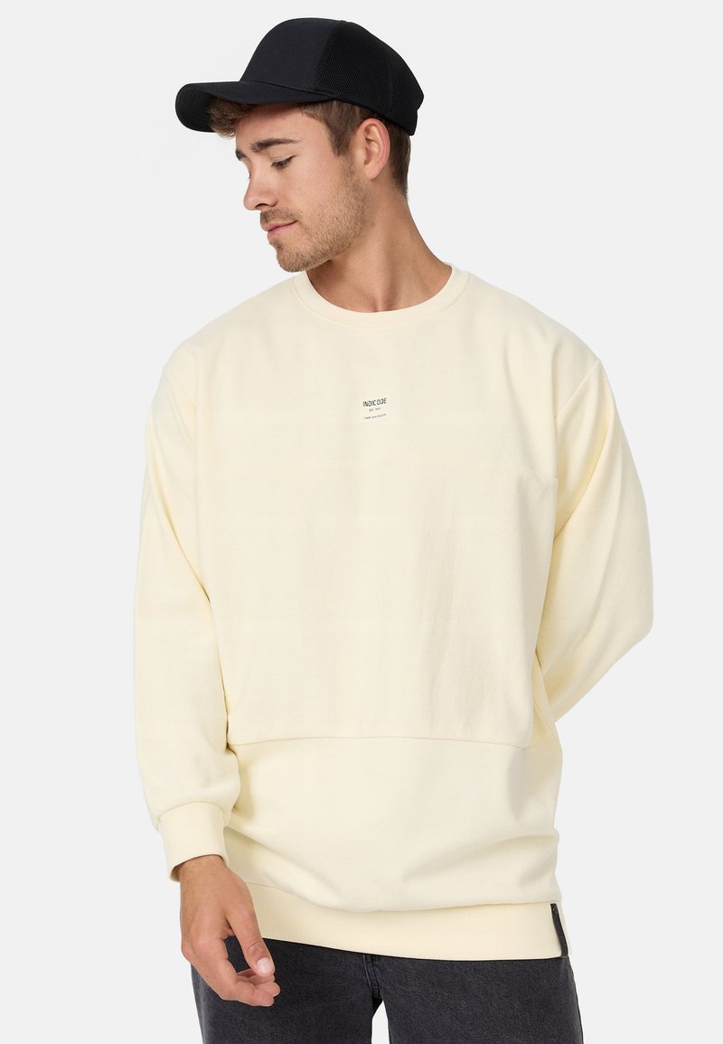 Cremfarbener Sweatshirt mit einer lockeren Passform, gerippten Bündchen und Saum sowie einem kleinen Markenlogo auf der Vorderseite. Kombiniert mit einer schwarzen Kappe.