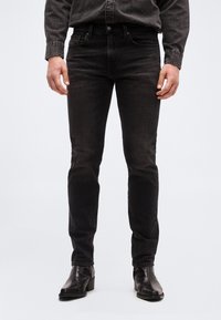 Άν δρας που φορά εφαρμοστά μαύρα τζιν και μαύρα δερμάτινα μποτάκια μέχρι τον αστράγαλο, στέκεται μπροστά σε ένα απλό φόντο.