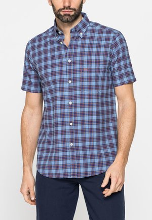 Uomo barbuto che indossa una camicia a maniche corte a quadri blu e rossi con pantaloni scuri, in piedi davanti a uno sfondo bianco.