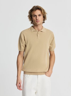 Giovane uomo con capelli biondi ricci indossa una polo beige a maniche corte e pantaloni bianchi, in piedi con una mano in tasca davanti a uno sfondo semplice.
