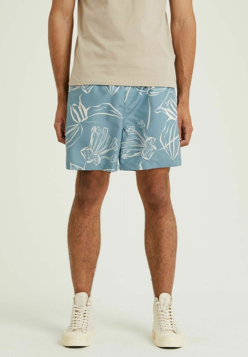 Homme portant une chemise beige, un short bleu à motifs floraux blancs et des baskets montantes blanches, debout devant un fond uni.
