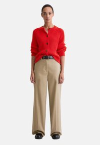 Czerwona prążkowana kardiganka z okrągłym dekoltem, trzema guzikami i skróconymi rękawami, zestawiona z beżowymi szerokimi spodniami i czarnymi butami.