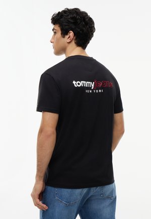 Joven con una camiseta negra "tommy jeans NEW YORK", mirando hacia la izquierda con la espalda a la cámara, combinado con vaqueros azules.