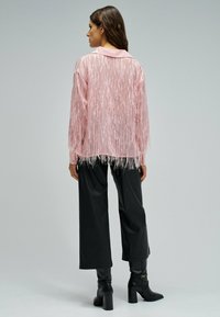 Femme vue de dos portant un chemisier rose transparent à franges, un pantalon large noir et des bottes à talons noires sur un fond uni.