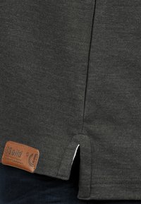 Mørkegrå T-shirt i tekstureret stof med en sideslids. Har et brunt læderlogo-patch ved kanten.