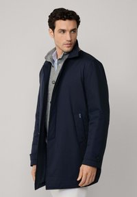 Marineblauwe waterdichte jas met een rits aan de voorkant, zijzakken en knoopmanchetten, gelaagd over een grijze trui en een blauwgestreepte blouse.
