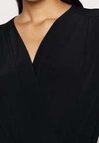 Abito aderente nero con scollatura a V profonda, realizzato in tessuto morbido. Presenta pieghe sottili sulle spalle e una silhouette elegante.