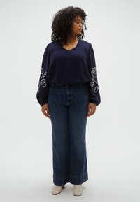 Γυναίκα με σγουρά μαλλιά που φοράει σκούρο μπλε μπλούζα με κεντήματα λουλουδιών στα μανίκια, τζιν παντελόνι φαρδύ στο κάτω μέρος και λευκά μποτάκια μέχρι τον αστράγαλο, στέκεται με κλειστά μάτια.