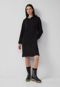 Schwarzes Kapuzenkleid mit langen Ärmeln, das einen lässigen Schnitt und seitliche Schlitze aufweist. Kombiniert mit klobigen schwarzen Plateaustiefeln aus gemischten Materialien.