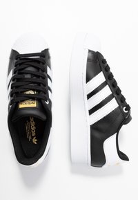 Mustat nahkasneakerit valkoisilla yksityiskohdilla, joissa on kolme raitaa, teksturoitu valkoinen kumikärki ja kultainen logotunnus kielekkeessä.