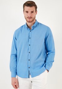 Camisa de manga larga azul clara con cuello camisero, cierre de botones en la parte delantera y botones oscuros en contraste. Textura suave, corte ajustado.