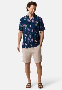 Korte mouwen, marineblauw button-up shirt met een tropisch palmprint in roze, geel en groen; gecombineerd met beige textured shorts en sandalen.