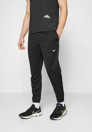 Persoon met zwart Nike Trail mouwloos shirt, zwarte Nike joggingbroek, witte sokken en zwarte Nike hardloopschoenen met neon accenten.