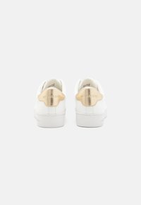 Baskets blanches avec un accent métallique doré au talon, présentant une finition en cuir lisse et une semelle en caoutchouc. Logo de la marque embossé sur le talon.
