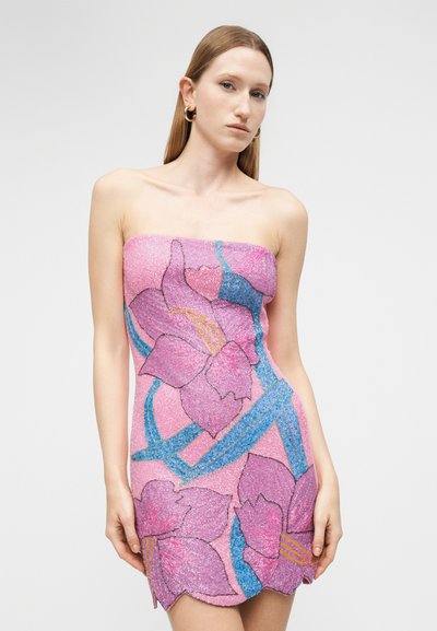 Ženska z ravnimi lasmi, oblečena v roza obleko brez naramnic z velikimi vijoličnimi cvetovi in modrimi listi na enobarvnem ozadju.