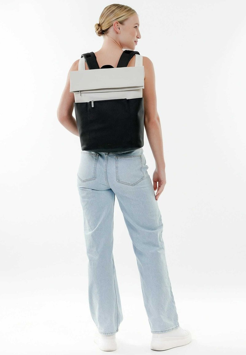 Mochila en blanco y negro con un diseño minimalista, forma rectangular, correas ajustables y un bolsillo frontal con cremallera. Textura suave.